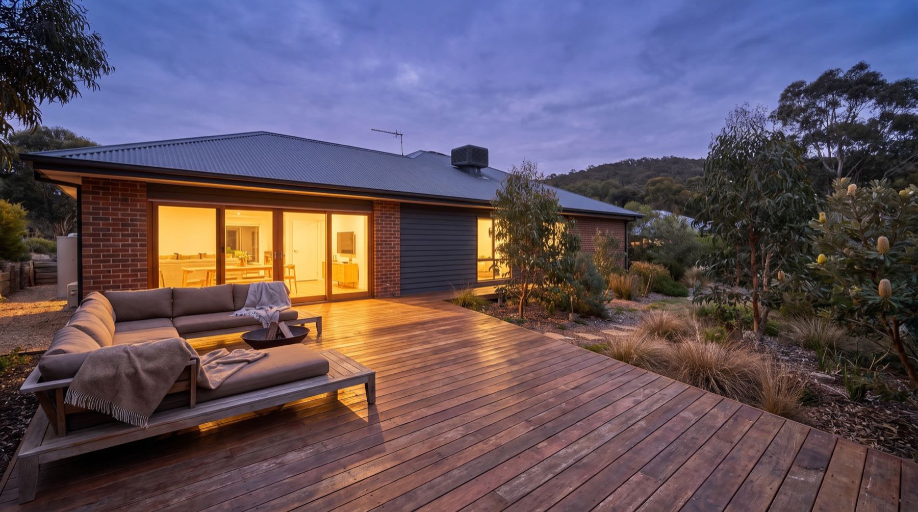 A finished timber deck and outdoor lounge lit from inside the house at dusk
